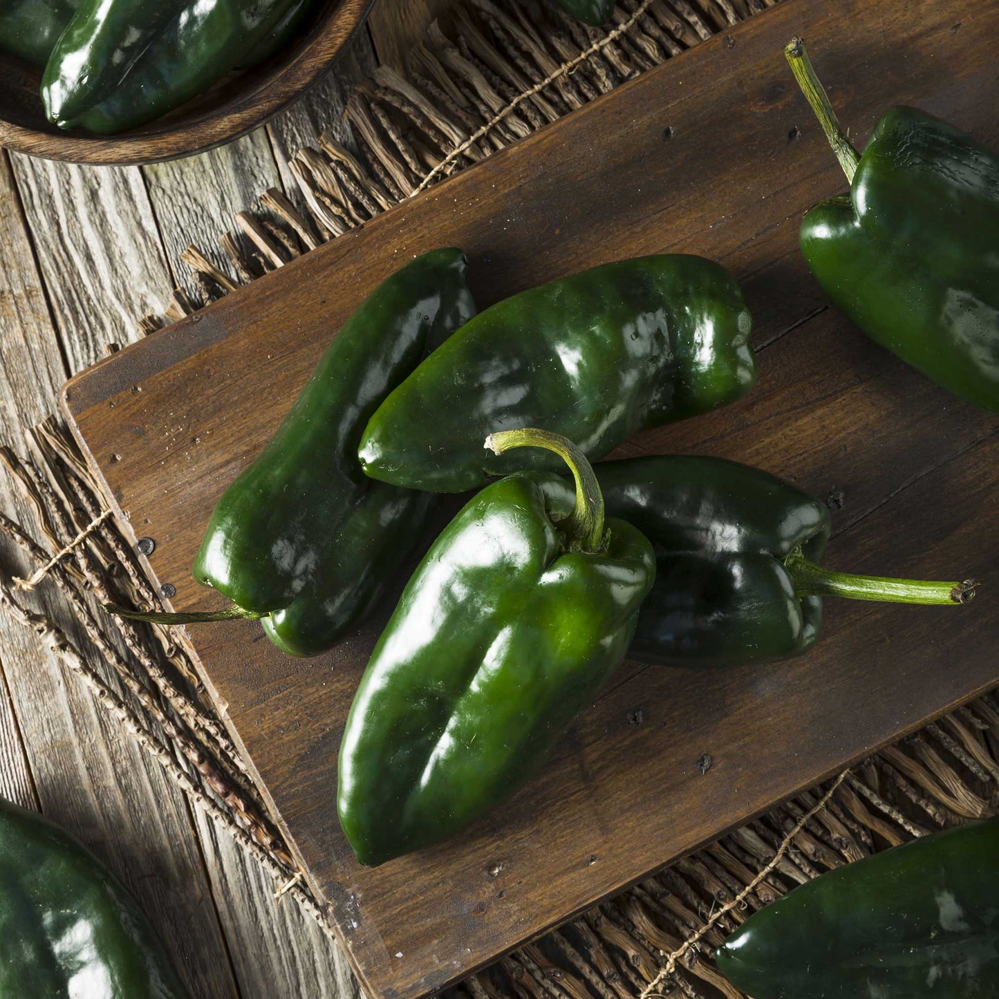 A pile of fresh, dark green Ancho or Poblano peppers on a wooden board.