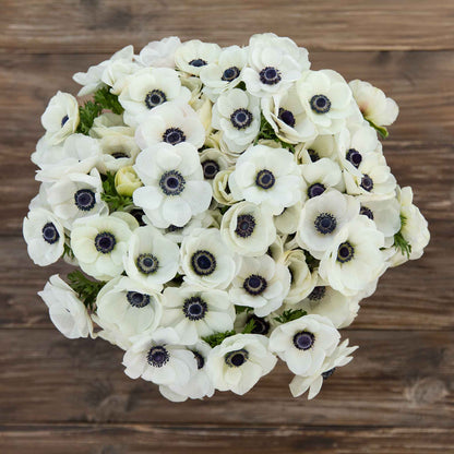 A boquet of white anemone flowers with dark centers and green foliage on a wooden table.