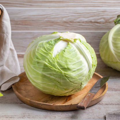 A round, green Brunswick cabbage head sits on a table, surrounded by large, dark green outer leaves.