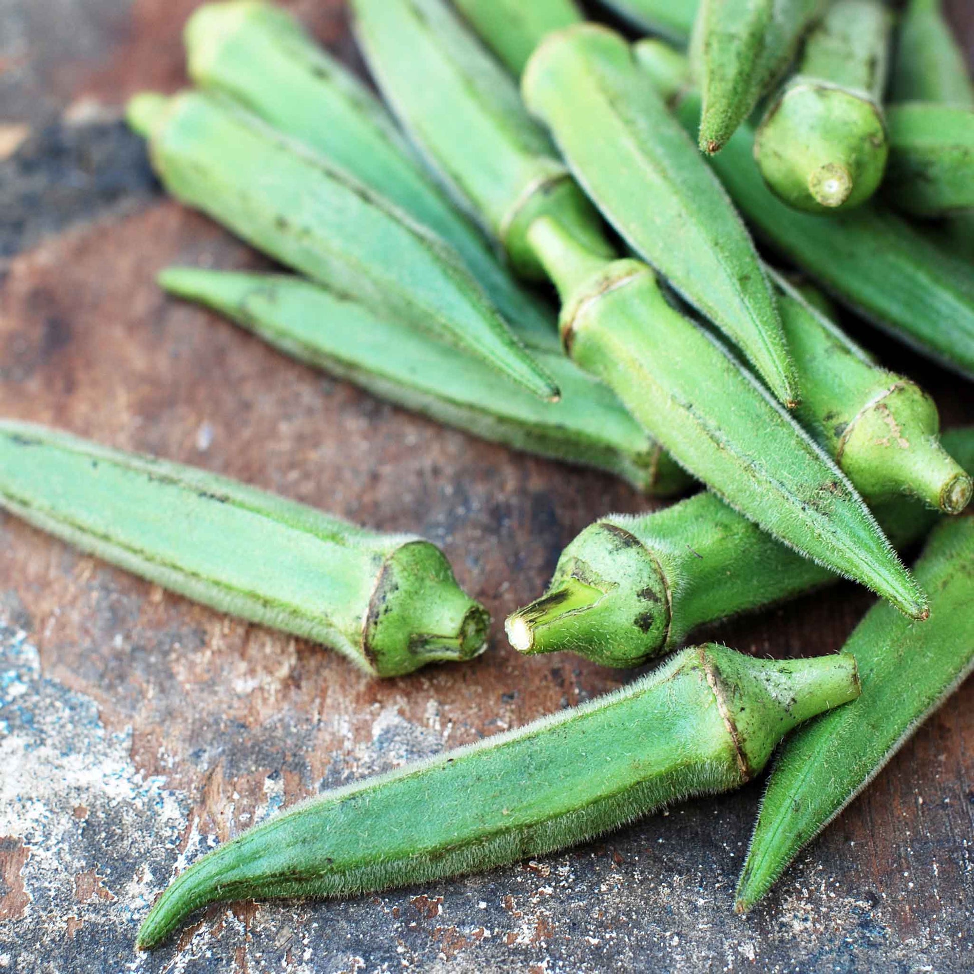 A close-up shot of vibrant green Emerald okra pods, freshly picked and arranged on a simple, rustic surface.