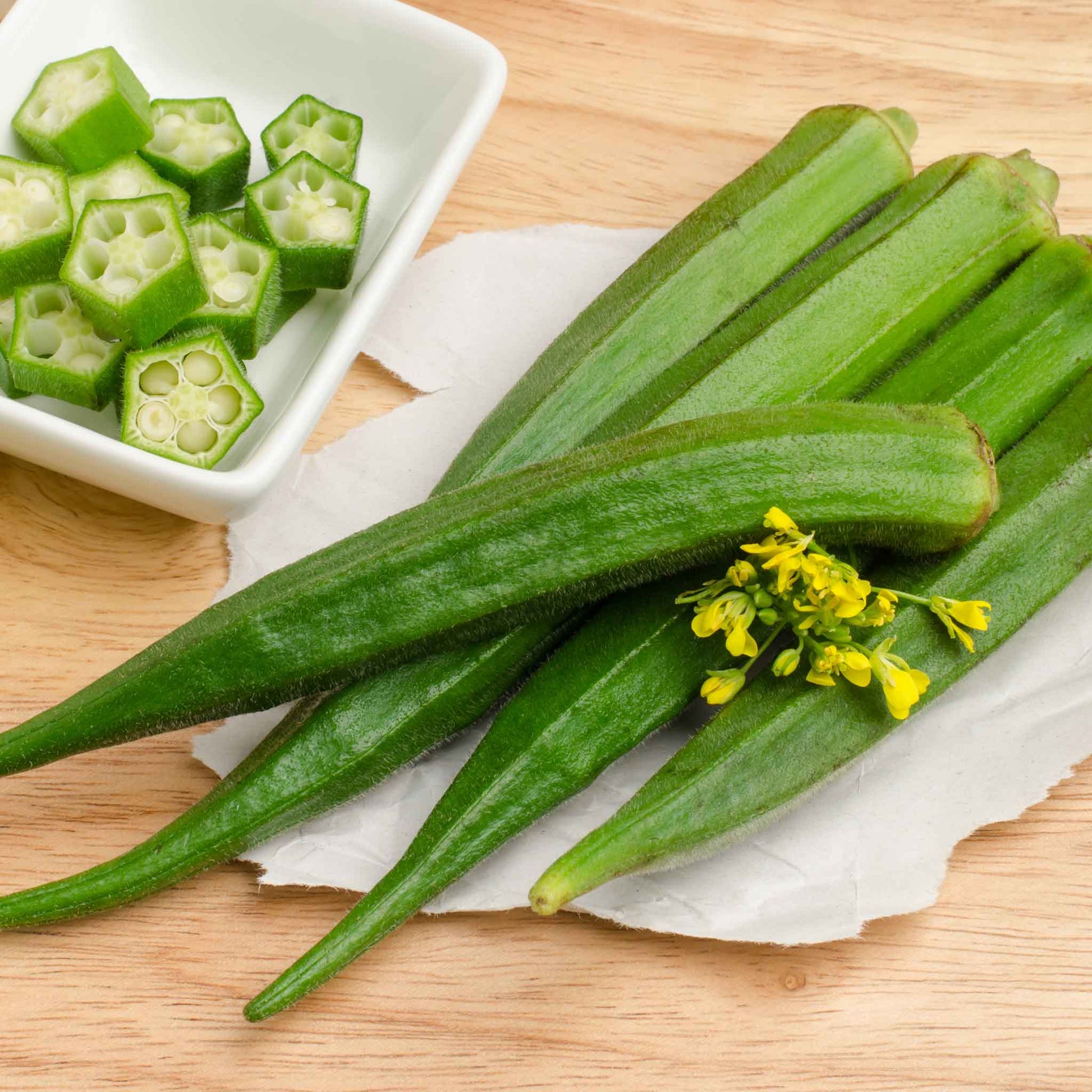 A close-up shot of green Emerald okra pods on a table with a bowl of sliced okra to the side.