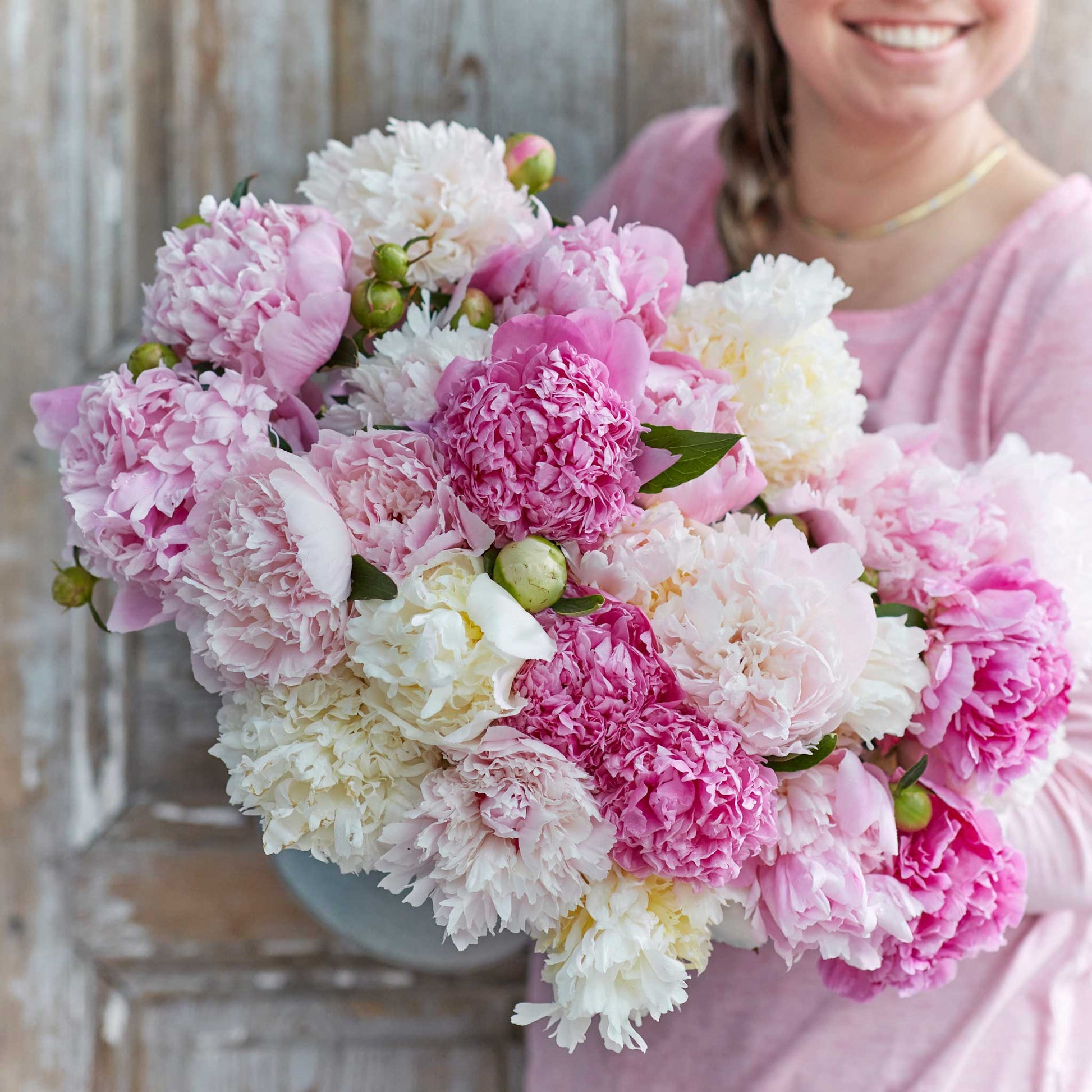 A person holding a bouquet of fluffy pink mix peony flowers with a wooden background.