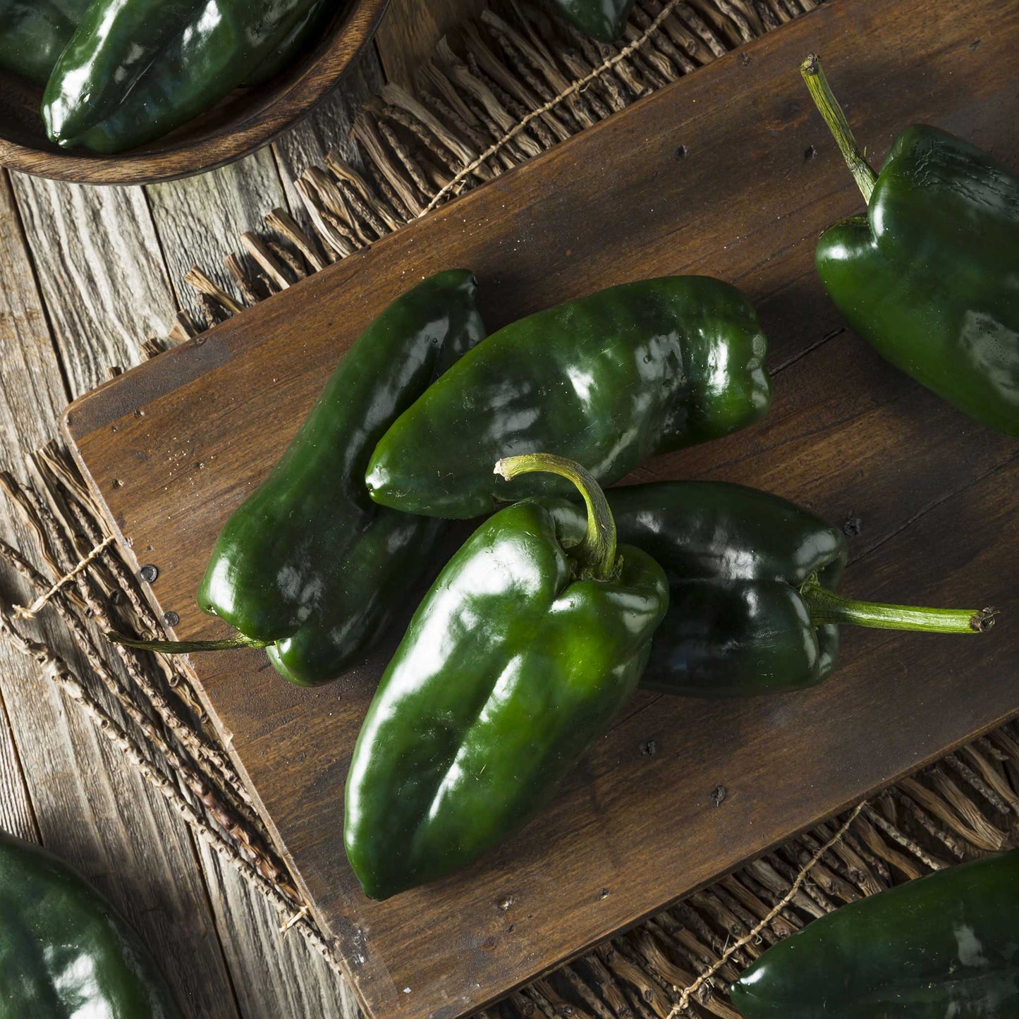 A pile of fresh, dark green Ancho or Poblano peppers on a wooden board.