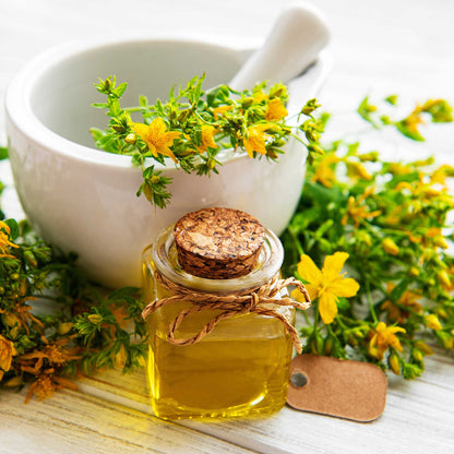 Small bottle of oil with cork lid, surrounded by st johns wort and a white mortar and pestle on a light wooden surface